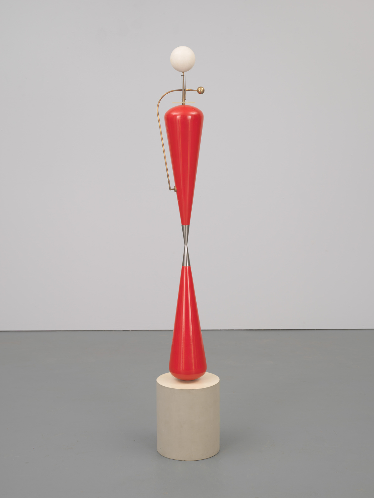 A modern sculpture featuring a red conical shape balanced on a cylindrical base, with a white sphere and a small golden sphere attached to a curved metal rod at the top. The background is a neutral gray with a slight shadow cast by the sculpture.