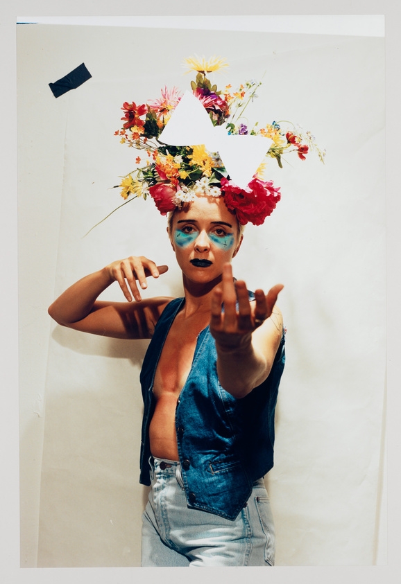 A person with a colorful floral headpiece and blue face paint reaching out towards the camera, wearing a denim vest and jeans, against a plain backdrop with a piece of black tape in the upper left corner.